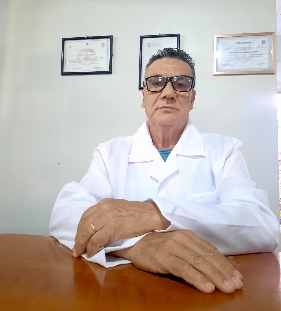 Dr. Leonel de jaleco branco e óculos, sentado à mesa de madeira, com três certificados emoldurados na parede ao fundo